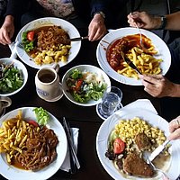 Gedeckter Tisch mit Tellern, die Nudeln und Gulasch enthalten. Desweiteren stehen Salatschüsslen und Bierkrüge auf dem Tisch. 