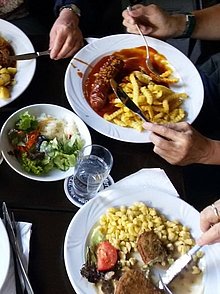 Gedeckter Tisch mit Tellern, die Nudeln und Gulasch enthalten. Desweiteren stehen Salatschüsslen und Bierkrüge auf dem Tisch. 