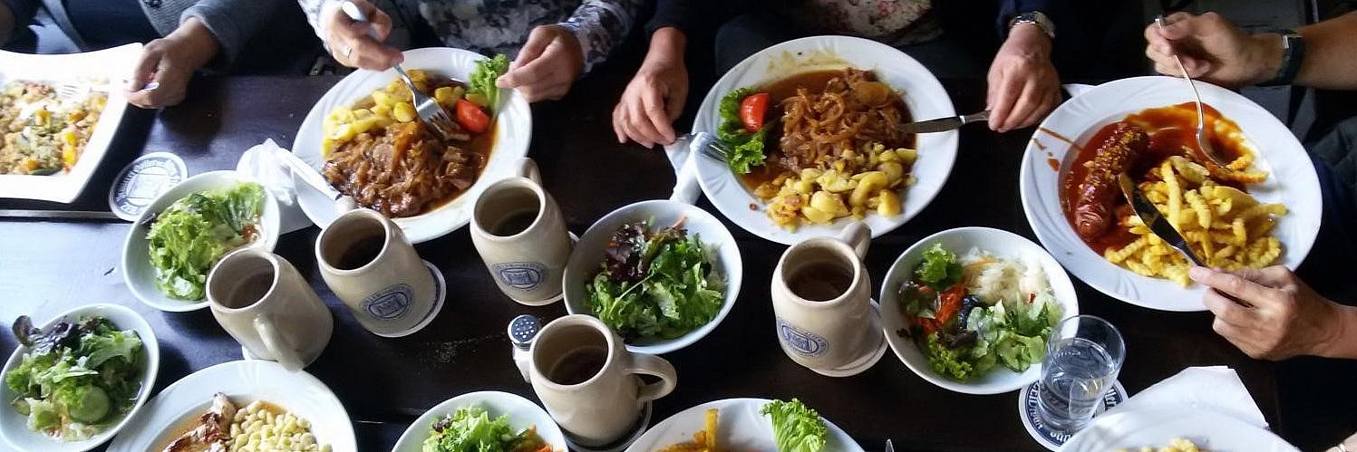 Gedeckter Tisch mit Tellern, die Nudeln und Gulasch enthalten. Desweiteren stehen Salatschüsslen und Bierkrüge auf dem Tisch. 
