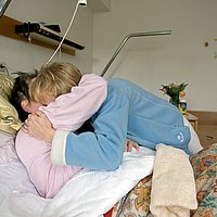 Eine Besucherin umarmt eine Frau im Krankenbett.