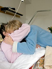 Eine Besucherin umarmt eine Frau im Krankenbett.