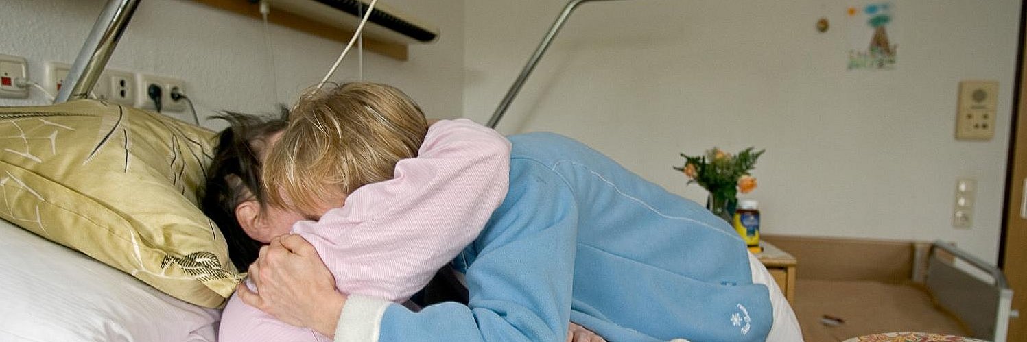Eine Besucherin umarmt eine Frau im Krankenbett.